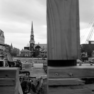 A black and white image of a construction site with heavy machinery in the foreground. In the background, there is a church with a tall spire and several older buildings. The scene is framed by a partially constructed wooden structure.