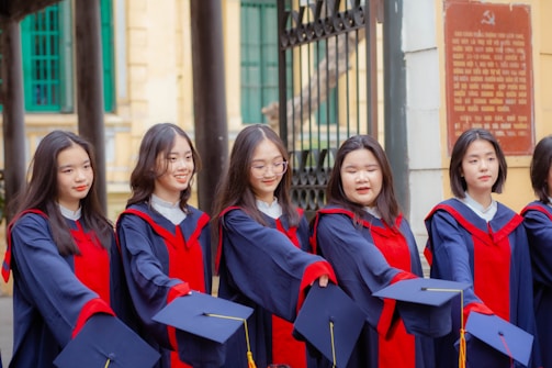 A group of young graduates wearing blue and red academic gowns and holding caps in their hands. They are standing side by side in a line, likely celebrating a graduation ceremony. The background features a building with green windows and a decorative gate.