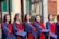 A group of young graduates wearing blue and red academic gowns and holding caps in their hands. They are standing side by side in a line, likely celebrating a graduation ceremony. The background features a building with green windows and a decorative gate.