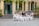 A group of people, mostly women dressed in white traditional dresses, are seated in front of a historic building with arched windows and green shutters. They sit on steps and chairs, some holding bouquets, appearing to enjoy a celebration or gathering.
