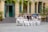A group of people, mostly women dressed in white traditional dresses, are seated in front of a historic building with arched windows and green shutters. They sit on steps and chairs, some holding bouquets, appearing to enjoy a celebration or gathering.