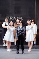 A group of six young people is standing in front of a store with the word 'PRADA' visible on the wall behind them. The central figure is wearing a black leather jacket, sunglasses, black pants, and boots. Surrounding them are five others wearing light-colored outfits, including dresses and casual wear.