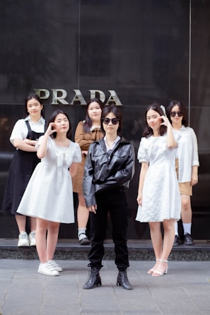 A group of six young people is standing in front of a store with the word 'PRADA' visible on the wall behind them. The central figure is wearing a black leather jacket, sunglasses, black pants, and boots. Surrounding them are five others wearing light-colored outfits, including dresses and casual wear.