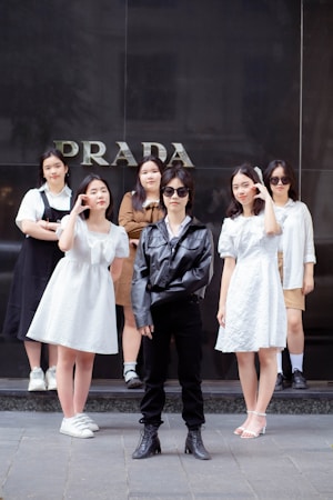 A group of six young people is standing in front of a store with the word 'PRADA' visible on the wall behind them. The central figure is wearing a black leather jacket, sunglasses, black pants, and boots. Surrounding them are five others wearing light-colored outfits, including dresses and casual wear.
