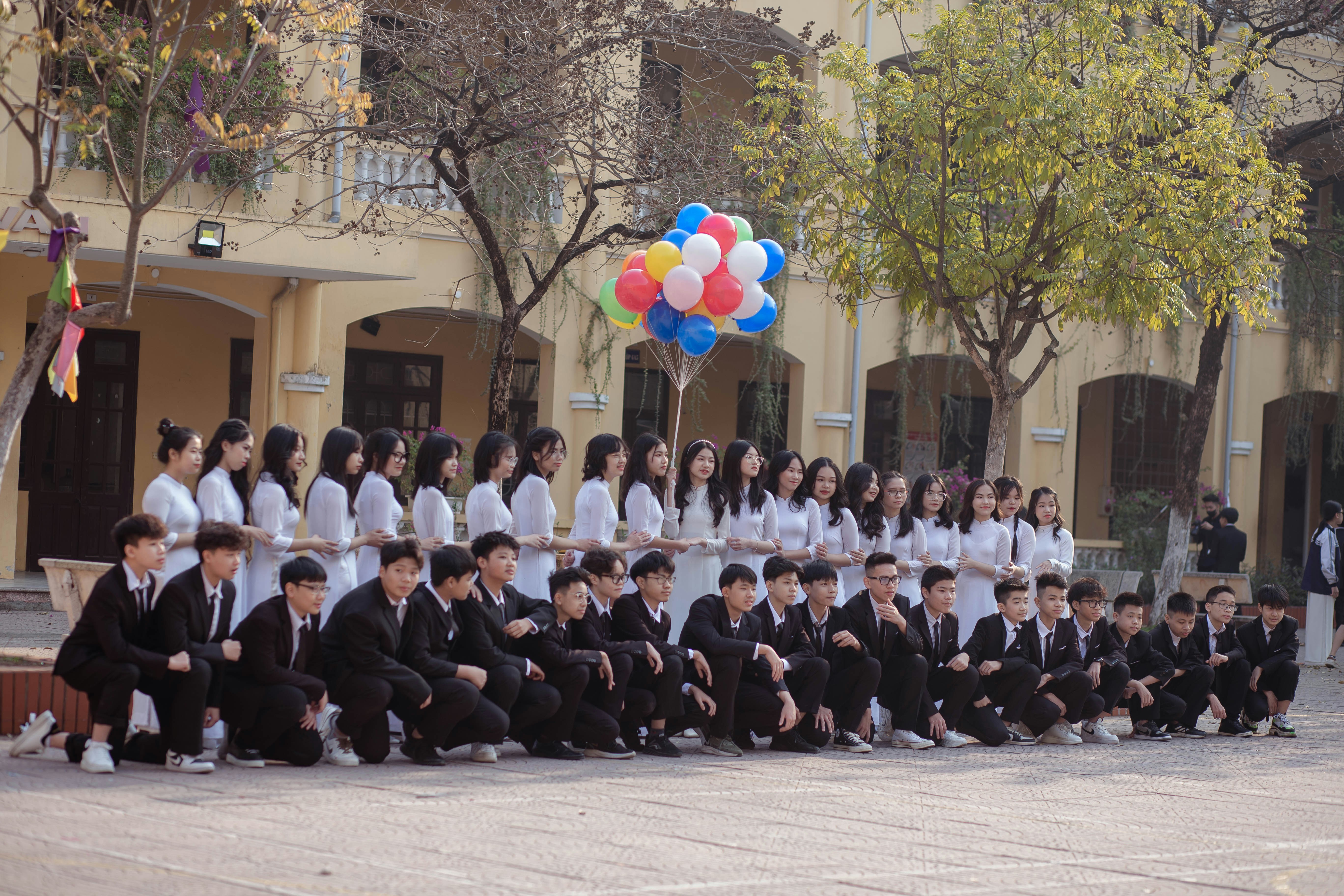 A group of students is arranged for a group photo in an outdoor setting. The girls are standing in a line wearing white dresses, while the boys are kneeling in front wearing black suits. One student holds a bunch of colorful balloons. The scene is set against a background of a yellow building with arched windows and some trees.