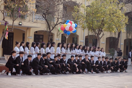 A group of students is arranged for a group photo in an outdoor setting. The girls are standing in a line wearing white dresses, while the boys are kneeling in front wearing black suits. One student holds a bunch of colorful balloons. The scene is set against a background of a yellow building with arched windows and some trees.