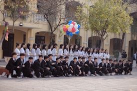 A group of students is arranged for a group photo in an outdoor setting. The girls are standing in a line wearing white dresses, while the boys are kneeling in front wearing black suits. One student holds a bunch of colorful balloons. The scene is set against a background of a yellow building with arched windows and some trees.