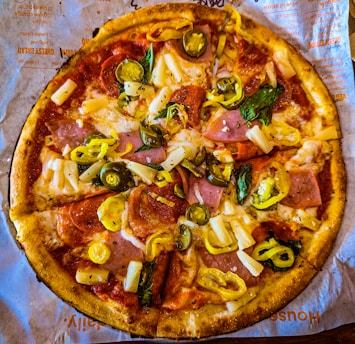 A pizza topped with slices of ham, pepperoni, pineapple chunks, jalapenos, green bell peppers, and melted cheese. It is placed on a piece of brown paper with writing on it.