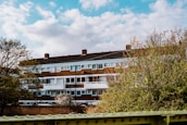 A mid-rise residential building with a brick facade and white balconies framed by trees in the foreground. The sky is partly cloudy, contributing to a serene and pleasant atmosphere.
