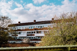 A mid-rise residential building with a brick facade and white balconies framed by trees in the foreground. The sky is partly cloudy, contributing to a serene and pleasant atmosphere.