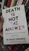 A book cover featuring bold text that reads 'DEATH IS NOT THE ANSWER' with a subtitle about understanding suicide and prevention. The author's name, Dr Anjali Chhabria, is displayed along with a mention of a foreword by Shabana Azmi. The background consists of a bookshelf with various other books.
