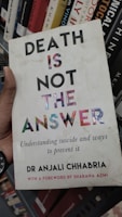 A book cover featuring bold text that reads 'DEATH IS NOT THE ANSWER' with a subtitle about understanding suicide and prevention. The author's name, Dr Anjali Chhabria, is displayed along with a mention of a foreword by Shabana Azmi. The background consists of a bookshelf with various other books.
