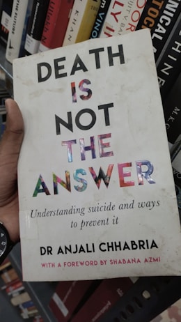 A book cover featuring bold text that reads 'DEATH IS NOT THE ANSWER' with a subtitle about understanding suicide and prevention. The author's name, Dr Anjali Chhabria, is displayed along with a mention of a foreword by Shabana Azmi. The background consists of a bookshelf with various other books.
