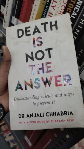 A book cover featuring bold text that reads 'DEATH IS NOT THE ANSWER' with a subtitle about understanding suicide and prevention. The author's name, Dr Anjali Chhabria, is displayed along with a mention of a foreword by Shabana Azmi. The background consists of a bookshelf with various other books.