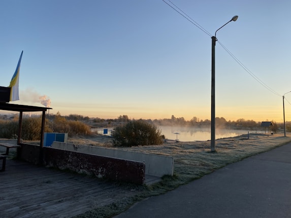 A serene early morning lakeside scene with a pathway stretching along the water. Bushes and trees are gently dusted with frost, and there is a subtle mist rising from the lake. The sky is clear and the horizon is lit with golden sunrise hues. A single streetlamp and a flag on a small structure are visible in the foreground.