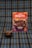 A package of 'Negrita' brand mazamorra morada flavor is placed on a plaid fabric background. In front of it, a bowl with dark purple dessert, presumably mazamorra morada, is garnished with two sticks of cinnamon.