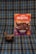 A package of 'Negrita' brand mazamorra morada flavor is placed on a plaid fabric background. In front of it, a bowl with dark purple dessert, presumably mazamorra morada, is garnished with two sticks of cinnamon.
