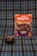 A package of 'Negrita' brand mazamorra morada flavor is placed on a plaid fabric background. In front of it, a bowl with dark purple dessert, presumably mazamorra morada, is garnished with two sticks of cinnamon.