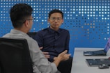 Two individuals are seated at a desk in discussion, each with a laptop open in front of them. The background features a blue wall with a pattern of diamond-shaped cutouts. One person is facing forward, while the other is turned slightly to the side.