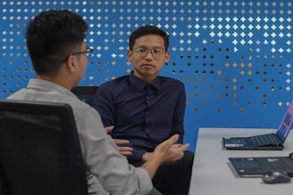 Two individuals are seated at a desk in discussion, each with a laptop open in front of them. The background features a blue wall with a pattern of diamond-shaped cutouts. One person is facing forward, while the other is turned slightly to the side.