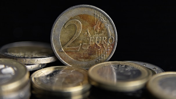 Several coins are scattered with a prominent two euro coin standing upright in the center. The coins have reflective metallic surfaces and intricate engravings featuring numbers and symbols.