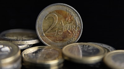 Several coins are scattered with a prominent two euro coin standing upright in the center. The coins have reflective metallic surfaces and intricate engravings featuring numbers and symbols.