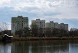 Several mid-rise residential buildings stand behind a line of leafless trees and a tranquil body of water. The buildings are primarily beige and green, with a cloudy sky above them. The water reflects the landscape, and there are reeds and a small structure located on the bank.
