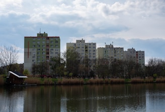 Several mid-rise residential buildings stand behind a line of leafless trees and a tranquil body of water. The buildings are primarily beige and green, with a cloudy sky above them. The water reflects the landscape, and there are reeds and a small structure located on the bank.