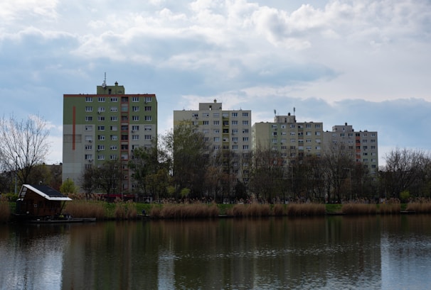 Several mid-rise residential buildings stand behind a line of leafless trees and a tranquil body of water. The buildings are primarily beige and green, with a cloudy sky above them. The water reflects the landscape, and there are reeds and a small structure located on the bank.