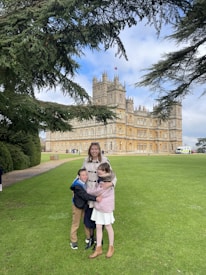 A woman and two children are standing on a green lawn in front of a large, historic castle-like building. They are positioned under the branches of a large tree which frames the scene. The sky is partly cloudy.