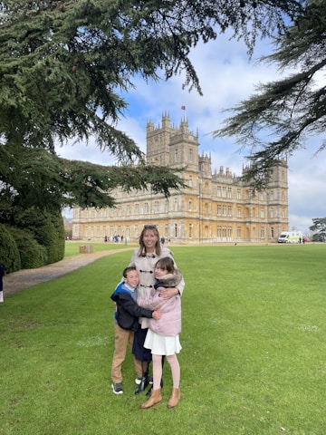 A woman and two children are standing on a green lawn in front of a large, historic castle-like building. They are positioned under the branches of a large tree which frames the scene. The sky is partly cloudy.