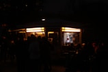 A small, illuminated kiosk is surrounded by people at night. The kiosk has bright yellow lights on the inside, revealing shelves lined with bottles and other items. Outside, silhouettes of people are visible, suggesting a gathering or social event.