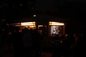 A small, illuminated kiosk is surrounded by people at night. The kiosk has bright yellow lights on the inside, revealing shelves lined with bottles and other items. Outside, silhouettes of people are visible, suggesting a gathering or social event.
