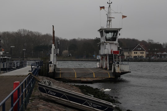 A docked ferry with a ramp is positioned on a waterway, surrounded by calm waters and flanked by a small boat named 'Fiete' moored on the rocky bank. The background features a row of trees and buildings, under a cloudy sky, creating a serene and slightly overcast atmosphere.
