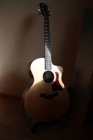 An acoustic guitar is positioned upright in a softly lit room, casting a shadow on the wall behind it. The light highlights the smooth wood grain of the guitar's body and neck, emphasizing its craftsmanship.