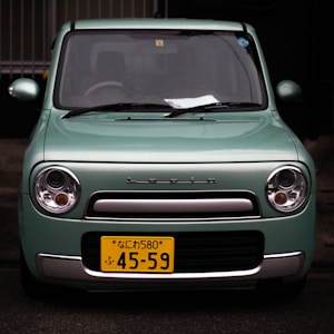 A mint green compact car with a front view, displaying a yellow Japanese license plate and the brand name 'Lapin' above the grille. The car has round headlights and a rectangular front grille with a silver trim. There are some papers visible on the dashboard inside the car.