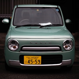 A mint green compact car with a front view, displaying a yellow Japanese license plate and the brand name 'Lapin' above the grille. The car has round headlights and a rectangular front grille with a silver trim. There are some papers visible on the dashboard inside the car.