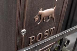 A metal post box features an embossed design of a cat holding an envelope, with the word 'POST' prominently displayed below. The box has a dark bronze color with a smooth, metallic finish and a keyhole lock.