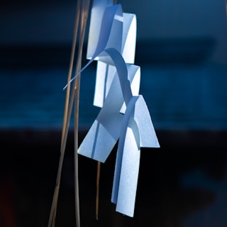 White paper strips are hanging vertically, possibly part of a traditional Shinto decoration, illuminated against a dark, blurred background. The paper appears carefully folded and arranged, casting soft shadows and highlighted by the natural light.