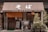 A traditional Japanese soba restaurant with wooden sliding doors and a brown fabric awning with Japanese characters. Several bicycles are parked in front, and potted plants are arranged beside the entrance. A chalkboard sign is positioned in front of the door.
