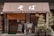 A traditional Japanese soba restaurant with wooden sliding doors and a brown fabric awning with Japanese characters. Several bicycles are parked in front, and potted plants are arranged beside the entrance. A chalkboard sign is positioned in front of the door.