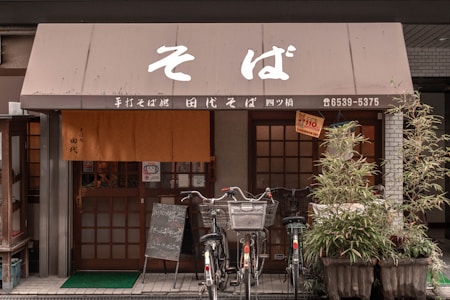 A traditional Japanese soba restaurant with wooden sliding doors and a brown fabric awning with Japanese characters. Several bicycles are parked in front, and potted plants are arranged beside the entrance. A chalkboard sign is positioned in front of the door.