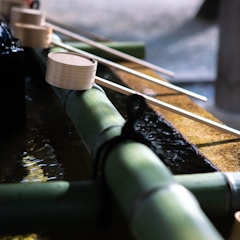 Ladles made of light wood are placed on top of a green bamboo water spout in a traditional setting. The bamboo is part of a water feature, possibly in a Japanese garden or temple, with gently flowing water underneath. Shadows and natural light create a serene and calm atmosphere.