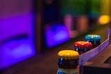 The image features a close-up view of colorful bottle caps lined up on a surface, with a background illuminated by soft, vibrant purple and blue lighting. The foreground caps are in sharp focus, showing intricate details, while the background is blurred, creating a depth effect.