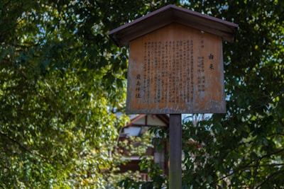 A weathered wooden sign with Japanese text stands amidst lush green foliage. The text appears to be historical or informational, possibly related to a nearby shrine or cultural site.