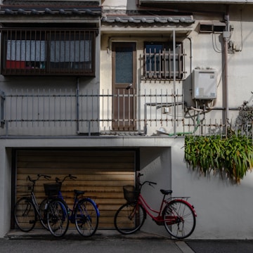 A residential building with a simple facade featuring an upper floor access door, small barred windows, and an external utility box. Three bicycles are parked in front of a closed garage door with a faded, rusted appearance. Green plants grow along a railing, adding a touch of nature to the urban setting.