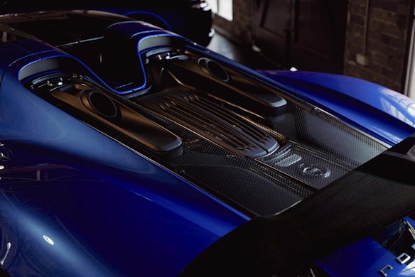 The image captures the rear section of a sleek, high-performance sports car with a focus on the engine and exhaust areas. The body features a glossy blue finish, complemented by carbon fiber detailing, providing a modern and luxurious feel.
