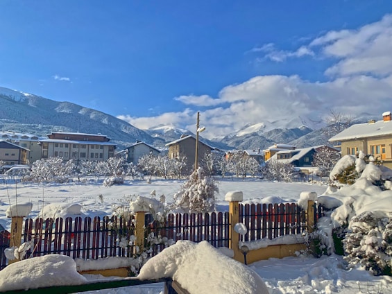 A snow-covered landscape features residential buildings and a fence in the foreground. Mountains dusted with snow are visible in the background under a clear blue sky. Leafless trees and shrubs are also blanketed with snow, adding to the wintry atmosphere.