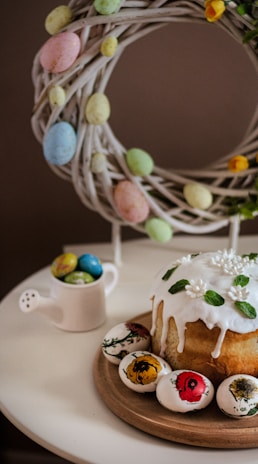 A decorative Easter scene featuring a braided wreath adorned with pastel-colored eggs hanging above a table. On the table, there is a round cake with white icing, topped with small flowers and green leaves. Next to the cake are several intricately painted Easter eggs with floral designs. A small white watering can is filled with additional colored eggs.