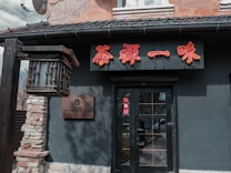 The exterior of a building features a stone pillar supporting a decorative lantern, and a black facade with red Chinese characters. A sign reads 'Chayna Town' next to a large entrance door.